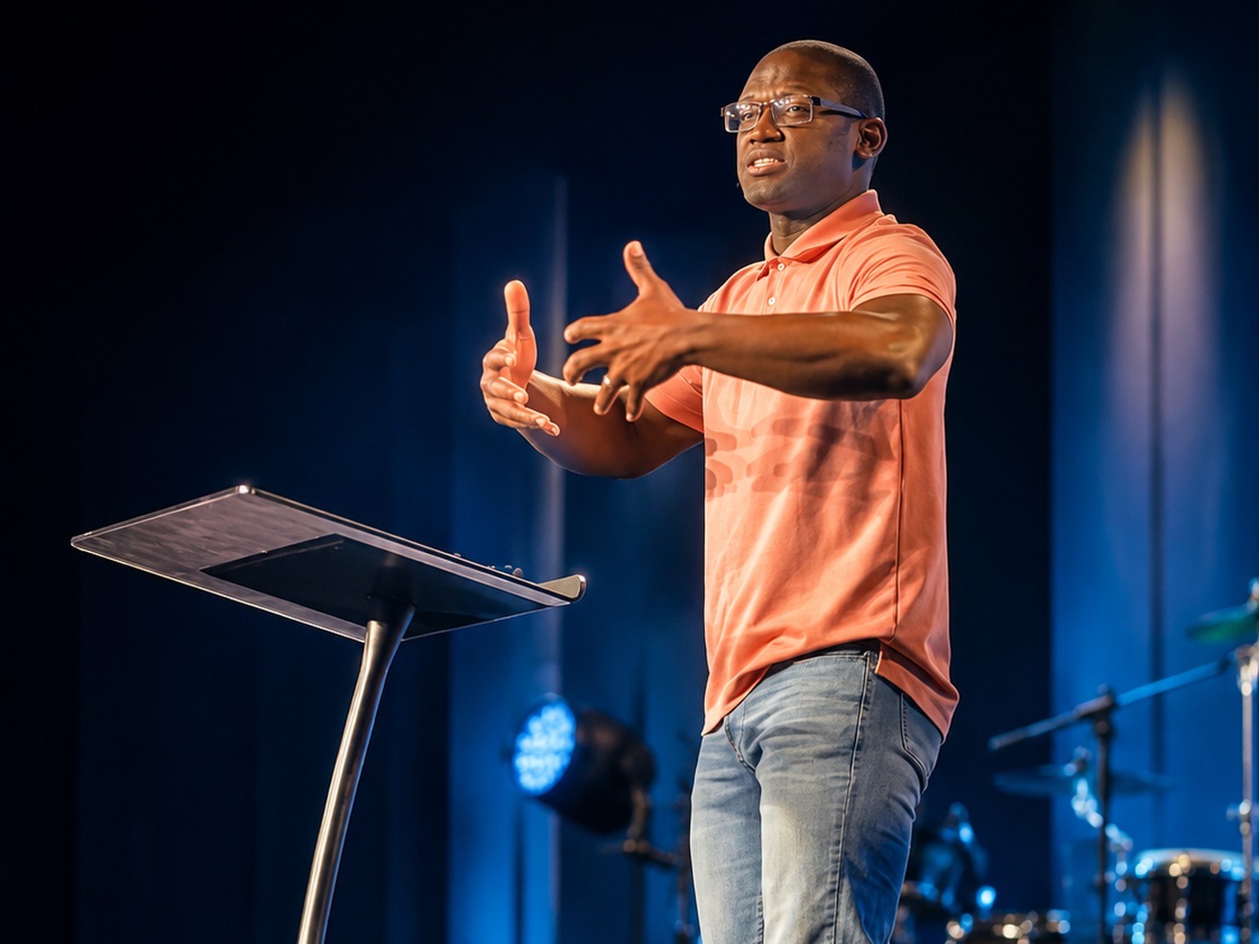 Denvil Lee teaching on stage in a coral polo, both hands reaching out as he makes a point.