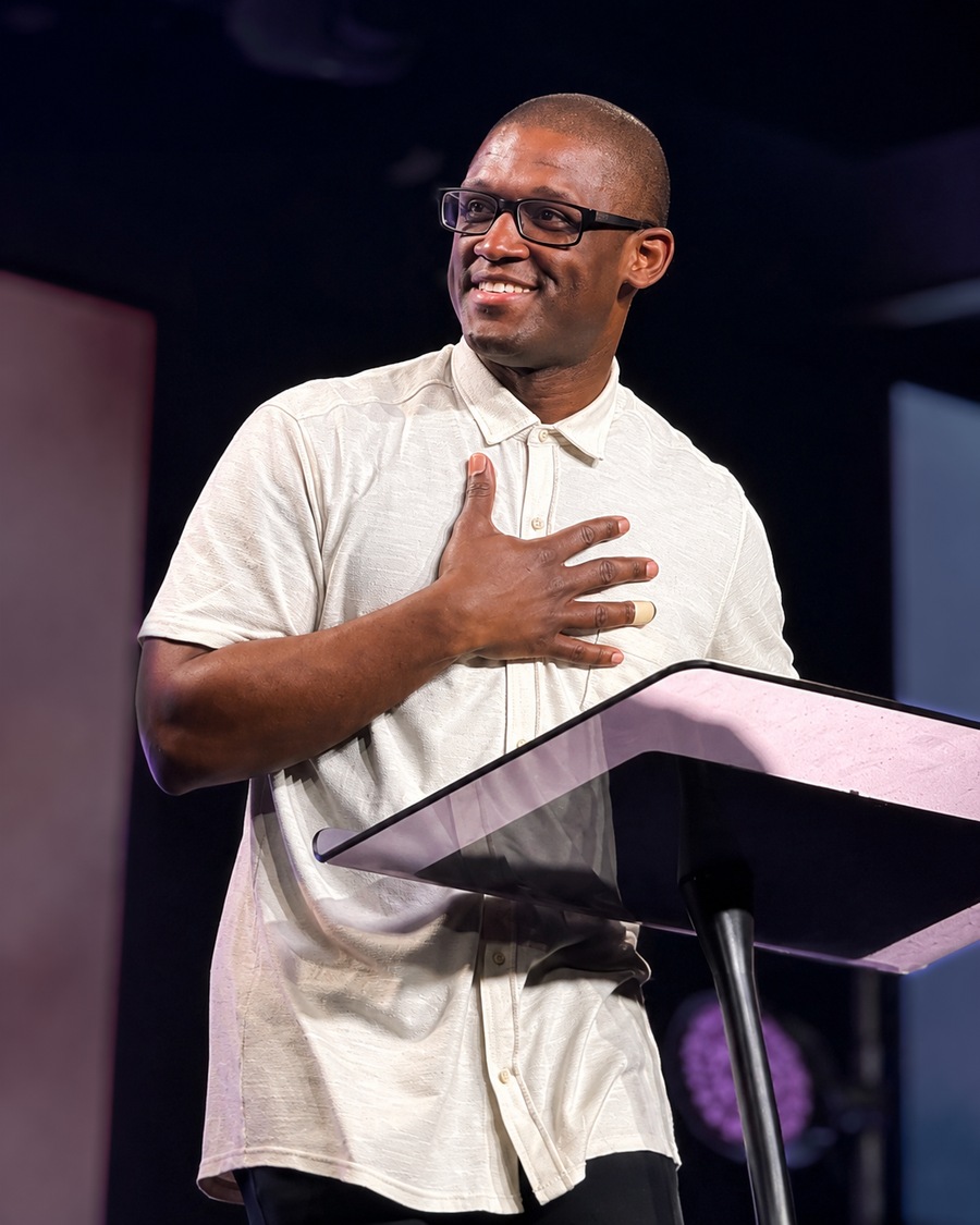 Denvil Lee at a lectern in a cream shirt, one hand resting on his chest.