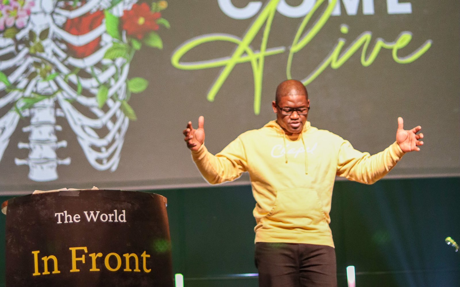 Denvil Lee teaching on stage in a yellow hoodie, arms open, under a Come Alive banner.